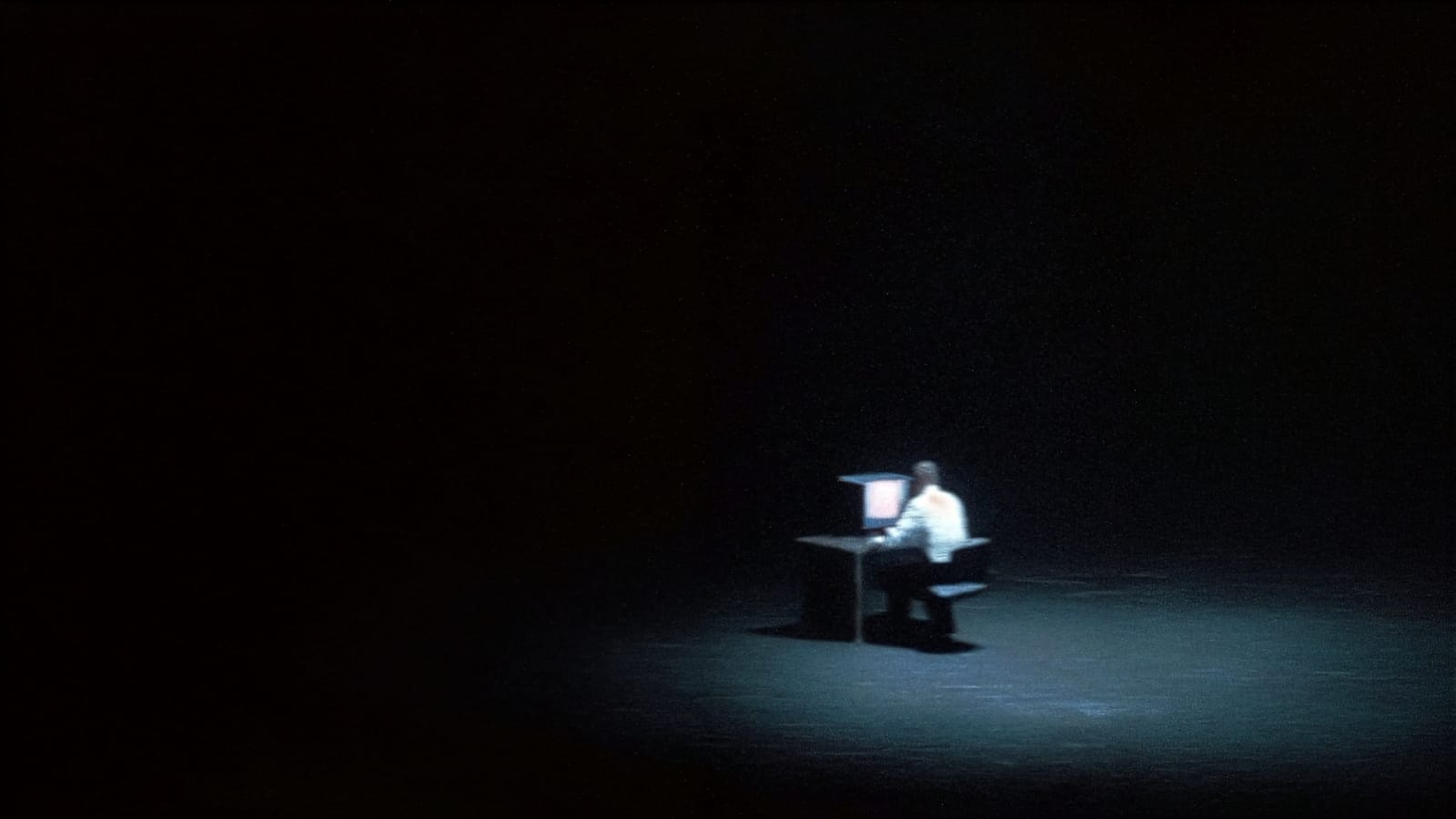 A lone developer at a desk under a single cold light, screens glowing in a black void.